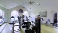 This fitness center is designed to support a comprehensive workout experience within a bright and welcoming environment. The space features soaring vaulted ceilings fitted with ceiling fans, promoting air circulation and comfort during exercise. Recessed lighting supplements the abundant natural light that pours in through a series of elegant arched windows, offering views of the surrounding exterior and creating an open, airy atmosphere throughout.

The facility is outfitted with a well-rounded selection of exercise equipment, including treadmills, elliptical machines, and a variety of strength training and resistance machines to accommodate a range of fitness goals. A wall-mounted flat-screen television provides entertainment for cardio sessions. The light-colored walls and pale flooring contribute to the clean, spacious feel of the room, while a decorative multi-panel artwork adds a touch of color to the otherwise neutral aesthetic.

The single-floor layout ensures easy accessibility, and the arrangement of equipment allows for comfortable movement throughout the space. Overall, this fitness center combines functional amenities with an inviting design, making it a well-suited environment for both casual and dedicated fitness routines.