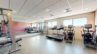 Located at 1331 Crossings Blvd in Shakopee, MN, this fitness center offers a well-equipped exercise room designed to support a range of workout routines. The space features a durable speckled floor, a drop tile ceiling with recessed and flush lighting, and a clean, functional layout that maximizes every square foot.

The cardio zone is anchored by multiple treadmills and an elliptical trainer, all equipped with individual digital displays. A full wall of mirrors runs along one side of the room, visually expanding the space while providing users with proper form feedback during workouts. Natural light filters in through a row of windows along the exterior wall, brightening the interior and offering outdoor views.

The strength training area includes a multi-function cable machine with a bench press station, a loaded weight rack with plates, and a freestanding dumbbell rack offering a graduated range of weights. A yoga or exercise mat rounds out the available accessories for stretching and floor exercises. A bold accent wall in deep red adds a dynamic visual element to the otherwise neutral color palette of whites and grays.

The room is accessed through a framed door and is designed to be efficient and user-friendly, making it a practical fitness amenity for residents or occupants of the building.
