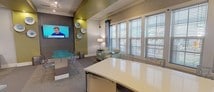 This single-floor community amenity space is thoughtfully designed with a contemporary aesthetic, blending functional areas into a cohesive, welcoming environment. The interior is characterized by a neutral palette of gray and warm beige, punctuated by bold olive-green ceiling accents that add visual interest throughout.

At the heart of the space is a curved reception desk with a sleek light-colored quartz countertop and rich wood veneer base, illuminated by striking cylindrical pendant lights. Adjacent to the reception area, a dedicated business center features multiple workstations equipped with desktop computers and ergonomic seating.

The lounge area offers comfortable seating arrangements alongside a wall-mounted flat-screen television and decorative plate wall art. Large windows dressed with horizontal blinds and teal accent curtains flood the space with natural light and provide views of the surrounding exterior.

A billiards area anchors the entertainment zone, centered around a full-sized pool table beneath a rectangular linear pendant fixture. The space transitions smoothly between tile and carpeted flooring, delineating each functional zone. A kitchenette with a curved countertop and undermount sink adds convenience, completing this well-appointed, multi-use gathering space.
