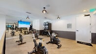 Located at 1464 Laurel Sound Ln in Sanford, Florida, this dedicated fitness center offers a thoughtfully designed workout environment within a generously sized single-floor space. The facility is accessed through sleek, floor-to-ceiling black-framed glass sliding doors, immediately establishing a modern, high-end aesthetic.

The interior features light hardwood-style flooring throughout, providing a clean and durable surface suited for a variety of exercise activities. Walls are finished in a contemporary two-tone scheme — crisp white on the upper half transitioning to a deep charcoal gray wainscoting below — lending the space a polished, intentional look. Pendant-style dome light fixtures with brushed metal finishes hang from the ceiling, complementing the overall design language.

The main workout area is outfitted with multiple Matrix stationary cycling bikes arranged for spin-style classes, as well as additional cardio equipment. A large wall-mounted display screen serves as a focal point, ideal for guided workouts or entertainment. A dedicated free-weight and stability ball station with a dark storage rack adds functional versatility.

Full-length mirrors along one wall enhance the sense of openness and allow users to monitor their form. A glass railing and open mezzanine-level overview further contribute to the airy, expansive feel of the space. The facility is well-ventilated with ceiling-integrated HVAC and includes clearly posted usage guidelines for residents.