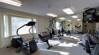 This single-floor fitness center offers a clean, functional space designed to support a well-rounded workout routine. The room is outfitted with a variety of exercise equipment, including a cardio stepper machine and a multi-station cable strength training system, providing options for both cardiovascular and resistance training. A weight bench adds further versatility to the workout area. Large wall-mounted mirrors span one side of the room, visually expanding the space and allowing users to monitor their form during exercise. Natural light streams in through multiple windows fitted with horizontal blinds, creating a bright and energizing atmosphere. The light-colored walls and neutral carpeted flooring contribute to a clean, open ambiance. Flush-mount, decorative ceiling light fixtures provide additional illumination, ensuring the space remains well-lit at all times. A wall-mounted HVAC unit ensures comfortable climate control year-round. The overall layout is thoughtfully arranged to maximize usable floor space while maintaining clear sightlines throughout the room, making it a practical and inviting environment for fitness activities.