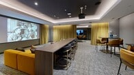 Located at 113 S Sixth St in downtown Minneapolis, this sophisticated multipurpose screening and lounge room offers a well-appointed single-floor layout designed for both entertainment and gathering. The space is anchored by a ceiling-mounted projector paired with a large projection screen, making it ideal for film screenings and presentations. Floor-to-ceiling gold velvet drapery lines the walls, adding warmth and acoustic control while lending a distinctly theatrical character to the room.

The interior is finished with dark, moody tones — charcoal-painted walls and a coffered black ceiling with integrated ambient LED lighting create an immersive atmosphere. Textured, patterned carpeting in grey and blue tones covers the floor throughout. A generous mustard-yellow sectional sofa with an assortment of decorative throw pillows anchors the screening area, providing comfortable viewing for a large group.

A long wooden bar counter with dark upholstered barstools serves as a transition zone between the screening and dining areas. The dining zone features a striking dark marble-topped table surrounded by cognac-colored upholstered chairs with brass-toned bases. Framed black-and-white photography and a bookshelf accent the walls, adding a curated, editorial feel. The overall design is cohesive, modern, and elegantly appointed for a premium communal experience.