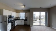 This single-floor residence offers a thoughtfully designed open-concept layout that seamlessly connects the kitchen and living areas. The kitchen is outfitted with sleek white cabinetry, dark stone countertops, and stainless steel appliances including a range, microwave, and dishwasher, all complemented by a undermount sink positioned beneath a window that allows for natural light. Track lighting on the ceiling provides additional illumination throughout the space. The flooring transitions from rich, dark hardwood in the kitchen and main living area to carpet near the living space, adding warmth and texture. A standout feature of this unit is the large sliding glass door that opens to a private balcony, filling the interior with abundant natural light and offering views of the surrounding neighborhood and skyline. The neutral, warm-toned walls throughout create a clean and versatile backdrop, while the open flow between the kitchen and living areas makes the space feel expansive and welcoming. The overall ambiance is contemporary and functional, well-suited for both everyday living and entertaining.