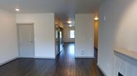 This single-floor space offers a straightforward and functional layout designed with everyday practicality in mind. The interior is defined by rich, dark hardwood flooring that runs throughout, creating a cohesive and grounded aesthetic from room to room. Clean white walls and flush ceiling light fixtures keep the space feeling bright and uncluttered, while warm accent walls add a subtle contrast that breaks up the neutral palette.

The kitchen is equipped with stainless steel appliances, including a full-sized refrigerator, and features cabinetry that makes efficient use of the available space. The open corridor-style layout allows for easy movement between the kitchen and the adjoining living areas, providing a natural flow throughout the unit.

A floor vent indicates a forced-air heating system, offering reliable climate control throughout the cooler months. The presence of a window at the far end of the hallway draws in natural light, brightening the interior and creating an inviting sense of depth. The overall ambiance is clean, modern, and move-in ready, with a no-frills design that prioritizes function and ease of use. This space is well-suited for those seeking a straightforward, well-maintained living environment with durable finishes and practical amenities.