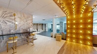Located at 15 Farm Springs Road in Farmington, Connecticut, this thoughtfully designed amenity space offers a generously sized, single-floor layout that seamlessly blends sophistication with functionality. The entrance area makes an immediate impression with polished marble-look flooring, a dramatic statement wall featuring a nature-inspired mural, and a sleek dark reception counter accented by gold bar stools and brass wall sconces. A striking wood-paneled ceiling lined with rows of exposed Edison-style bulbs adds warmth and visual interest overhead. The space transitions into an open lounge area furnished with clean-lined white seating and arched niche details, bathed in recessed lighting. A glass-enclosed conference room with full-height glazing offers a private yet visually connected meeting area, equipped with a long table and ample seating. Beyond the lounge, the space opens into a flexible recreation and event area featuring large-format gray tile flooring, a billiards table as a central focal point, and abundant natural light streaming through oversized windows. A more open, versatile flex area with light tile flooring completes the layout, suitable for gatherings or additional programming. Throughout, the design maintains a cohesive contemporary aesthetic with neutral tones, gold accents, and a polished, professional ambiance.