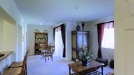 This single-floor space presents a traditionally styled interior characterized by warm, classic furnishings and a cohesive, lived-in aesthetic. The main living area features plush wall-to-wall carpeting in a neutral tone, contributing to a soft and comfortable ambiance throughout. Ceilings are smooth and painted in a crisp white, lending a clean, open feel to the room.

Natural light filters in through multiple windows dressed with rich, dark burgundy curtains accented by decorative valances, adding depth and a touch of elegance to the space. A substantial antique-style wooden armoire serves as a prominent focal point along one wall, providing both visual weight and practical storage. Nearby, a wooden coffee table anchors the seating area, complemented by a wicker accent chair.

A charming bistro-style dining set with ornate wrought-iron chairs occupies one corner, creating an intimate nook. Adjacent to it, a green-painted hutch displays decorative dishware and collectibles, reinforcing the traditional character of the space. Floral arrangements and decorative accents are thoughtfully placed throughout, enhancing the warm, welcoming atmosphere of this classically appointed interior.