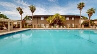 Located at 3800 Michael Blvd in Mobile, Alabama, this outdoor pool area is a well-appointed communal amenity space designed for relaxation and recreation. The centerpiece is a generously sized rectangular swimming pool with striking blue tile trim and crystal-clear turquoise water, offering an inviting atmosphere year-round in the mild Gulf Coast climate.

The pool is surrounded by a broad, paved deck constructed with attractive interlocking pavers, providing plenty of room for lounging and socializing. Multiple chaise lounge chairs are positioned throughout the deck area, offering residents comfortable spaces to relax both in the sun and in the shade.

A prominent white pergola structure anchors one end of the pool deck, providing a shaded gathering area with additional seating. The space is enclosed by decorative iron fencing accented with brick columns, lending a sense of privacy and security while maintaining an open, airy feel.

Tall palm trees and mature lush greenery frame the surroundings, enhancing the tropical ambiance characteristic of coastal Alabama. The multi-story residential buildings visible in the background feature a combination of neutral earth tones, pitched rooflines, and brick accents that complement the outdoor setting. The pool area is well-lit for evening use and offers a welcoming, resort-like environment for residents of the community.