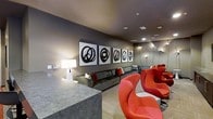 This single-floor lounge space showcases a carefully curated contemporary design that balances bold contrasts with a sophisticated, cohesive palette. The room is anchored by a series of four large-scale framed abstract artworks mounted along a deep charcoal grey wall — each piece featuring circular black brushstroke motifs on white backgrounds, creating a dramatic and cohesive gallery-style display.

Recessed ceiling lighting provides ambient illumination throughout the space, complemented by wall-mounted sconces and a classic white table lamp that adds a warm glow near the seating area. The grey sectional sofa, dressed with red and neutral accent pillows, runs along the back wall and provides generous seating.

A cluster of sculptural, curved red lounge chairs positioned in the foreground serves as a striking focal point, injecting energy and personality into the space. Their organic forms contrast with the clean, angular lines of a stone or concrete reception-style counter visible in the lower left corner. The flooring features a subtly patterned light-toned carpet, softening the overall atmosphere while tying together the room's modern, design-forward aesthetic.