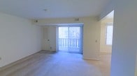 This single-floor space features a spacious, open living area finished with light-colored carpeting and neutral beige walls that create a clean, understated atmosphere. The room is anchored by a large sliding glass door dressed with vertical blinds, which floods the interior with natural light and provides direct access to an outdoor balcony, offering views of the surrounding exterior. A secondary window on the adjacent wall further enhances the brightness of the space. A louvered door, likely leading to a closet or utility area, adds a functional element to the room. HVAC vents are positioned along the ceiling, ensuring efficient climate control throughout. The ceiling is smooth and white, complementing the overall light and airy feel of the interior. The open layout allows for flexible furniture arrangement, making the living area adaptable to a variety of uses. The combination of natural light, neutral finishes, and direct outdoor access gives the space a practical yet comfortable character suitable for everyday living.