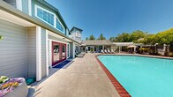 Located at Bay Village in Santa Rosa, California, this multi-level amenity facility serves as the community's central gathering hub. The main floor offers a generous footprint, while an upper level provides additional functional space. The architectural design is highlighted by soaring vaulted ceilings with exposed white beams, flooding the interiors with natural light through clerestory windows and large glass panes throughout.

The welcoming lounge area features rich hardwood flooring, a bold statement fireplace with a crisp white surround, wall-mounted television, and stylish pendant lighting. Comfortable seating arrangements create an inviting atmosphere for relaxation. Adjacent to the lounge, a well-appointed coffee bar showcases dark countertops, white cabinetry, a chalkboard accent wall, and pendant lighting, providing a cozy refreshment station.

The fitness center is spacious and well-equipped, featuring a full suite of cardio machines including treadmills and ellipticals, a cable weight machine, a dumbbell rack, and a workout bench. Floor-to-ceiling mirrors amplify the sense of space, and large windows offer views of the surrounding greenery.

Outside, a large sparkling pool serves as the centerpiece of the outdoor amenity area, bordered by an expansive concrete deck lined with lounge chairs. A pergola-shaded seating area and lush mature trees complete the resort-style atmosphere.