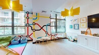 Located at 1 Fineran Way, this single-floor children's playroom is a thoughtfully designed, energetic space built to inspire creativity and active play. The most striking design feature is the bold, large-scale transit map mural that wraps across the walls, rendered in vivid red, blue, yellow, and black — immediately capturing the imagination upon entry. Light hardwood flooring runs throughout the main area, providing a clean and durable surface that complements the lively décor. Overhead, sculptural yellow pendant light fixtures add a playful architectural element to the bright white ceiling. Large windows along the perimeter flood the room with natural light and offer views of the surrounding outdoor courtyard, creating an open and airy atmosphere. The space is divided into several distinct activity zones: a soft-mat area with interlocking red and blue foam tiles houses a wooden play structure complete with a slide, ideal for active play. A dedicated seating area features child-sized tables and modern chairs for creative activities. A built-in white cabinetry unit anchors a media wall with a large flat-screen television and toy storage. Additional features include a play kitchen, a cozy navy-blue nook with a lime green cushioned bench, and colorful framed artwork displayed throughout.
