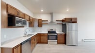 This single-floor unit offers a thoughtfully designed layout that maximizes its compact footprint. Light wood-tone flooring runs continuously throughout the space, creating a cohesive and airy aesthetic. The open-plan kitchen and living area forms the heart of the unit, with the kitchen standing out as a particularly well-appointed space. Warm-toned wood cabinetry—both upper and lower—is paired with crisp white countertops and a chevron-patterned tile backsplash. A stainless steel appliance suite includes a refrigerator, electric range with a wall-mounted hood, built-in microwave, and dishwasher, offering full functionality. Recessed lighting illuminates the kitchen, while open shelving adds a modern touch.

The spacious bedroom features large windows that frame urban views, a ceiling fan, and baseboard heating. A closet and an adjacent door connect conveniently back toward the kitchen area. The bathroom is clean and straightforward, featuring a walk-in shower, toilet, and wood-look flooring that mirrors the rest of the unit. Neutral warm-beige walls throughout lend the space a calm, modern feel suited to everyday living.