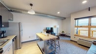 Located at 3659 SE 29th Avenue in Portland, Oregon, this single-floor unit presents a thoughtfully designed interior with a contemporary aesthetic. The open-concept kitchen and living area serves as the heart of the space, where exposed cylindrical ductwork lends an industrial character to the otherwise clean, neutral palette. The kitchen is equipped with stainless steel appliances, dark cabinetry, a white subway tile backsplash, and a deep sink. A pendant light fixture illuminates a butcher-block island on casters, which doubles as a casual dining surface with seating for several. The adjoining living area features wood-look flooring transitioning to carpet, recessed lighting, and a window with wood-trim framing that invites natural light and exterior views, complemented by a wall-mounted HVAC unit. The bedroom is a calm, well-proportioned retreat finished with wood-look flooring, a ceiling fan with integrated light, and a sliding closet. The full bathroom includes a tiled tub-shower combination, a floating vanity with a vessel sink, and wood-look tile flooring, all finished in a clean, modern style consistent with the rest of the unit.