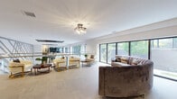Located at 22 Sussex in Hackensack, New Jersey, this thoughtfully designed amenity floor offers a seamless blend of comfort, productivity, and contemporary style. The open-plan layout is unified by large-format light stone tile flooring and crisp white walls, creating a bright, airy atmosphere throughout.

The spacious lounge area serves as a focal point, featuring a curved velvet sofa, gold-accented armchairs, and a round coffee table arranged beneath an elegant multi-bulb chandelier. Floor-to-ceiling sliding glass doors and wide-frame windows frame lush greenery outside, flooding the interior with natural light and providing a refreshing connection to the outdoors.

A dedicated entertainment and dining zone includes a rustic wood dining table with bench seating, wall-mounted flat-screen televisions, and a striking gold accent table, anchored by a deep charcoal feature wall that adds bold contrast to the otherwise neutral palette.

The co-working area is equipped with adjustable standing desks, desktop computers, and task lighting, offering a functional workspace within the communal setting. Decorative mirrors and framed artwork add personality to this zone.

A glass-railed staircase and decorative metalwork railing provide architectural interest, while potted greenery is thoughtfully placed throughout to enliven the space. The overall ambiance is modern, welcoming, and versatile.
