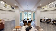 This thoughtfully designed single-floor community space is organized into several distinct functional areas, each with its own character while maintaining a cohesive aesthetic throughout. The overall layout is open and expansive, with notably high ceilings that are accentuated by a wide upper display ledge showcasing large-scale curated artwork, adding a gallery-like quality to the interior.

The central dining area serves as a visual focal point, anchored by a substantial dark wood dining table with upholstered seating beneath a sleek pendant light. The floor beneath transitions into a striking geometric patterned tile in muted blue, gray, and white tones, creating a defined zone within the space. Floor-to-ceiling glass sliding doors at the far end open directly to an outdoor courtyard, flooding the area with natural light and providing seamless indoor-outdoor connectivity.

Adjacent to the dining zone, a comfortable lounge area features a built-in wood media wall with integrated shelving, cabinetry, and a large mounted flat-screen television. Mixed seating including a cognac leather sofa, velvet accent chairs, and modern lounge chairs are arranged around a central coffee table atop a colorful area rug.

A separate meeting or conference nook offers a dramatic black marble table surrounded by cane-back chairs, set within a refined wainscoted wall enclosure. Dark wood-look tile flooring and white panel wainscoting unify the space throughout.