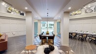 This thoughtfully designed single-floor community space is organized into several distinct functional areas, each with its own character while maintaining a cohesive aesthetic throughout. The overall layout is open and expansive, with notably high ceilings that are accentuated by a wide upper display ledge showcasing large-scale curated artwork, adding a gallery-like quality to the interior.

The central dining area serves as a visual focal point, anchored by a substantial dark wood dining table with upholstered seating beneath a sleek pendant light. The floor beneath transitions into a striking geometric patterned tile in muted blue, gray, and white tones, creating a defined zone within the space. Floor-to-ceiling glass sliding doors at the far end open directly to an outdoor courtyard, flooding the area with natural light and providing seamless indoor-outdoor connectivity.

Adjacent to the dining zone, a comfortable lounge area features a built-in wood media wall with integrated shelving, cabinetry, and a large mounted flat-screen television. Mixed seating including a cognac leather sofa, velvet accent chairs, and modern lounge chairs are arranged around a central coffee table atop a colorful area rug.

A separate meeting or conference nook offers a dramatic black marble table surrounded by cane-back chairs, set within a refined wainscoted wall enclosure. Dark wood-look tile flooring and white panel wainscoting unify the space throughout.