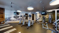 Located at 5877 N Nevada Ave in Colorado Springs, Colorado, this fitness center is a thoughtfully designed, single-floor facility built to support a comprehensive workout experience. The space is anchored by a bold athletic aesthetic, most notably a large-scale running track mural that spans a prominent wall, setting an energetic and motivating tone throughout.

The cardio zone features a row of modern treadmills equipped with individual screens, positioned along windows that allow natural light to fill the space. Alongside these are elliptical cross-trainers and stair-climbing machines, offering a variety of low- and high-impact options. The strength training area is well-stocked with a full rack of dumbbells, cable machines, a functional trainer, and a weight bench, catering to users of all fitness levels.

Overhead, large circular pendant lights provide bright, even illumination across the rubber-floored workout areas. Multiple wall-mounted flat-screen TVs are positioned throughout the space for entertainment during workouts. Accent walls feature a combination of reclaimed wood paneling and graphic typography, adding a contemporary, design-forward character to the facility.

A corridor with polished concrete flooring connects ancillary rooms, including what appears to be restroom or utility access. Ceiling fans contribute to air circulation, ensuring comfort during intense training sessions. The overall design balances function and style, creating an inviting environment for daily fitness routines.