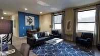 This well-appointed single-floor space makes a strong visual impression through its thoughtful use of color, light, and design. The living area serves as the centerpiece, anchored by a striking cobalt blue accent wall adorned with a vibrant fish-themed art piece that adds a dynamic focal point to the room. A charcoal tufted sofa, a coordinating blue armchair, and a sleek metallic coffee table are arranged over a bold navy geometric area rug, creating a cohesive and contemporary aesthetic.

Natural light filters in through two generously sized windows dressed with elegant striped curtains, illuminating the warm beige tones of the surrounding walls and light wood-style flooring. Recessed ceiling lighting complements the natural light, ensuring the space remains bright and inviting throughout the day.

Transitioning seamlessly from the living area, the dining space is visible in the background, featuring a wooden dining table and chairs alongside lush potted greenery that brings a touch of nature indoors. A classic floor lamp adds warmth to the corner of the room, while decorative wall art and carefully chosen accessories throughout reflect a refined, modern sensibility. The overall ambiance is stylish, comfortable, and welcoming.