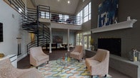 The clubhouse at River Oak Apartments is an architecturally striking communal space defined by its dramatic double-height ceilings and open, airy layout. A sweeping spiral staircase with sleek black metal railings serves as the centerpiece, connecting the main level to an open upper mezzanine overlooking the lounge below. Large multi-pane windows rise toward the ceiling on multiple walls, flooding the interior with natural light and offering views of the surrounding outdoor areas.

The main lounge area is anchored by a tile-surround fireplace framed by a white mantel and a bold abstract artwork above, adding a refined focal point to the space. Comfortable upholstered seating arranged around a glass-top coffee table rests atop a colorful geometric area rug, creating an inviting gathering area. French doors adjacent to the fireplace open directly to an outdoor patio and pool area, seamlessly connecting indoor and outdoor living.

A dedicated game room features a pool table and shuffleboard table beneath the soaring windows, offering recreational options bathed in natural light. Colorful artwork is displayed throughout, contributing to the space's contemporary, well-curated aesthetic. The overall ambiance is sophisticated yet approachable, designed to serve as a welcoming hub for the community.