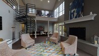 The clubhouse at River Oak Apartments is an architecturally striking communal space defined by its dramatic double-height ceilings and open, airy layout. A sweeping spiral staircase with sleek black metal railings serves as the centerpiece, connecting the main level to an open upper mezzanine overlooking the lounge below. Large multi-pane windows rise toward the ceiling on multiple walls, flooding the interior with natural light and offering views of the surrounding outdoor areas.

The main lounge area is anchored by a tile-surround fireplace framed by a white mantel and a bold abstract artwork above, adding a refined focal point to the space. Comfortable upholstered seating arranged around a glass-top coffee table rests atop a colorful geometric area rug, creating an inviting gathering area. French doors adjacent to the fireplace open directly to an outdoor patio and pool area, seamlessly connecting indoor and outdoor living.

A dedicated game room features a pool table and shuffleboard table beneath the soaring windows, offering recreational options bathed in natural light. Colorful artwork is displayed throughout, contributing to the space's contemporary, well-curated aesthetic. The overall ambiance is sophisticated yet approachable, designed to serve as a welcoming hub for the community.