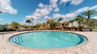 This outdoor amenity space centers around a generously sized, curved swimming pool with crystal-clear blue water, set within a well-maintained residential community that evokes a distinctly Florida tropical aesthetic. The pool is flanked by a broad, paved deck finished in warm-toned interlocking stone tiles, providing ample space for lounge seating and relaxation. Depth markers are clearly visible along the pool's edge, indicating a shallow, leisure-friendly design accessible to a wide range of residents and guests.

Lush landscaping frames the entire area, with tall swaying palm trees, vibrant red-leafed plants, and mature tropical greenery creating a resort-like atmosphere beneath an open, sun-filled sky. The surrounding residential buildings, clad in earth-toned and green exteriors, are visible in the background, reinforcing the cohesive character of the community.

Adjacent to the pool deck stands a single-story clubhouse or amenity building with a peach-toned stucco exterior, a light metal roof, and large glass French doors that open directly onto the patio. Wrought iron outdoor furniture is positioned along the deck, inviting residents to gather and socialize in a comfortable, shaded setting. The overall ambiance is bright, open, and welcoming, reflecting a laid-back outdoor lifestyle suited to warm-weather living.