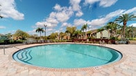 This outdoor amenity space centers around a generously sized, curved swimming pool with crystal-clear blue water, set within a well-maintained residential community that evokes a distinctly Florida tropical aesthetic. The pool is flanked by a broad, paved deck finished in warm-toned interlocking stone tiles, providing ample space for lounge seating and relaxation. Depth markers are clearly visible along the pool's edge, indicating a shallow, leisure-friendly design accessible to a wide range of residents and guests.

Lush landscaping frames the entire area, with tall swaying palm trees, vibrant red-leafed plants, and mature tropical greenery creating a resort-like atmosphere beneath an open, sun-filled sky. The surrounding residential buildings, clad in earth-toned and green exteriors, are visible in the background, reinforcing the cohesive character of the community.

Adjacent to the pool deck stands a single-story clubhouse or amenity building with a peach-toned stucco exterior, a light metal roof, and large glass French doors that open directly onto the patio. Wrought iron outdoor furniture is positioned along the deck, inviting residents to gather and socialize in a comfortable, shaded setting. The overall ambiance is bright, open, and welcoming, reflecting a laid-back outdoor lifestyle suited to warm-weather living.