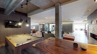 This single-floor amenity space showcases a sophisticated blend of industrial and contemporary design. Exposed concrete columns and a raw concrete ceiling contrast beautifully with warm wood accents, creating a dynamic yet inviting atmosphere. At the forefront, a well-appointed billiards table sits beneath sleek pendant lighting, complemented by a striking live-edge wood communal table that serves as a natural centerpiece. The open-plan layout flows seamlessly into a comfortable lounge area furnished with modern armchairs and low-profile seating, including rich leather ottomans that add a refined touch. Recessed lighting brightens the interior, while the space extends into a visible fitness area in the background, equipped with exercise machines and separated by glass partitions that maintain visual openness. The overall design balances function and aesthetics, with carefully curated furnishings, mixed textures, and a neutral color palette anchored by warm wood tones and cool concrete finishes. The generous ceiling height enhances the sense of openness, making this a versatile, well-designed communal space suited for both recreation and relaxation.