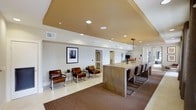 The WiFi Lounge is a thoughtfully designed communal space that blends functionality with modern aesthetics. The open-plan layout is defined by a warm, two-tone ceiling detail — a recessed golden-tan tray ceiling that draws the eye upward and adds architectural depth to the room, complemented by recessed lighting throughout.

A prominent bar-height counter with a wood-paneled base runs along one side of the space, accompanied by sleek chrome-framed bar stools upholstered in rich brown leather. A modern linear pendant light hangs above the counter, adding an elegant focal point. A fresh floral arrangement provides a welcoming touch of color to the surface.

On the opposite side, a casual seating area features matching brown leather chairs with chrome legs, arranged alongside compact accent tables — ideal for individual work or small group gatherings. A framed black-and-white photograph adorns the wall, adding a refined artistic element to the lounge.

The flooring transitions from light-toned tile to a rich brown area rug, subtly defining the seating zones. Neutral white walls keep the space feeling bright and open, while HVAC vents and a clean architectural layout ensure year-round comfort. Additional rooms are accessible through a doorway at the rear, extending the utility of this well-appointed lounge.