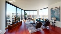 This contemporary urban residence showcases an open, sophisticated design defined by dramatic floor-to-ceiling glass walls that flood every room with natural light and frame sweeping city skyline and neighborhood views. Rich, deep-toned hardwood floors run throughout the main living areas, anchoring the space with warmth and visual continuity.

The expansive living room is a standout, with wraparound corner glazing offering panoramic urban vistas and ample space for comfortable seating arrangements. Adjacent to the living area, the kitchen is outfitted with warm wood cabinetry, stainless steel appliances including a large refrigerator and professional-grade range hood, a central island with bar seating, and track lighting overhead — combining style with full functionality.

The primary bedroom is generously proportioned, featuring large windows with city views, plush carpeting, and a serene, restful atmosphere. A second bedroom offers its own oversized window with open outdoor views, providing a bright and private retreat. Both bedrooms are served by well-appointed bathrooms — one featuring a sleek glass-enclosed walk-in shower with clean white tile finishes, and the other a full bath with mosaic tile flooring and a wall-mounted vanity.

A dedicated laundry area with stacked washer and dryer units and open shelving rounds out this well-equipped, modern residence.