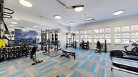 Located at 4106 Brickell Drive in Fairfax, VA, this fitness center offers a generously sized, single-floor exercise room designed to support a comprehensive workout experience. The space features a clean, modern aesthetic with white walls, patterned carpet tiles in grey and vivid blue, and flush-mounted ceiling lights complemented by dual ceiling fans that promote air circulation throughout.

The exercise room is divided into distinct functional zones. The strength training area includes a power rack with weight plates, a cable machine system, a full dumbbell rack, and adjustable benches, all positioned alongside floor-to-ceiling mirrors that amplify the sense of space. A designated cardio section houses multiple treadmills and additional cardio machines, including a stationary bike and elliptical. An accessories station offers medicine balls, resistance bands, and a foam roller rack.

A striking motivational mural covering one full wall serves as a bold visual centerpiece, reinforcing an energetic atmosphere. A wall-mounted television provides entertainment during workouts. A large grid of windows along one exterior wall floods the room with natural light and offers views of the tree-lined surroundings, enhancing the overall ambiance. The combination of ample equipment, thoughtful layout, and bright, open design makes this fitness center a highly functional and inviting space.