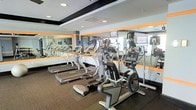 This fitness center offers a clean, modern workout space designed for effective cardio training. The room is lined with large floor-to-ceiling mirrors framed in warm orange accents, visually expanding the space and allowing users to monitor their form during exercise. Recessed ceiling lighting provides consistent, bright illumination throughout the room, complementing the overall sleek aesthetic. The equipment lineup includes multiple treadmills and elliptical cross-trainers, each unit outfitted with individual display screens for entertainment or workout tracking. The machines are arranged in a well-organized layout, allowing comfortable spacing between each unit. The flooring features a durable, textured carpet in a neutral tone, providing both comfort underfoot and a degree of noise reduction. The mirrored walls reflect natural light from windows visible to one side, contributing to an open, airy atmosphere. The overall design balances functionality with a polished, contemporary look, making this fitness center a practical and inviting space for daily cardio workouts.