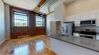 Located at 548 Atwells Ave in Providence's vibrant Federal Hill neighborhood, this single-floor loft-style unit showcases a compelling blend of industrial heritage and contemporary updates. The living area immediately stands out with its exposed brick walls, original hardwood floors, and wooden ceiling beams that speak to the building's historic character. Soaring ceilings amplify the sense of openness, while large multi-pane factory-style windows flood the space with abundant natural light and offer views of the surrounding neighborhood. A ceiling fan with an integrated light fixture adds both function and style to the main living area, which also features a wall-mounted mini-split system for climate control.

The kitchen is thoughtfully designed with white shaker cabinetry reaching toward the high ceilings, dark granite-style countertops, and a full suite of stainless steel appliances, including a refrigerator, electric range, and microwave. A peninsula with a built-in sink provides additional prep space and casual seating potential. Exposed ductwork running along the ceiling reinforces the industrial aesthetic throughout. The bathroom continues the warm hardwood flooring and features clean, neutral finishes with tiled shower surround.