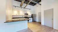 Located in Rochester's vibrant East End neighborhood, this one-bedroom, one-bathroom urban loft at 50 Prince Street blends industrial character with modern finishes. Soaring ceilings with exposed ductwork and bold steel beams define the open-concept living space, giving the unit a distinctive loft aesthetic throughout. Light hardwood flooring flows seamlessly from room to room, enhancing the bright, airy feel.

The kitchen is a standout feature, offering white shaker cabinetry, dark granite countertops, a central island with a built-in sink, and a full suite of stainless steel appliances including a range, over-the-range microwave, and refrigerator. Track lighting illuminates the space with a modern, industrial touch. The kitchen opens directly to the combined dining and living area, making it ideal for entertaining.

The bedroom is generously proportioned and benefits from a dramatic bank of tall, narrow windows that flood the room with natural light and offer views of the surrounding Rochester streetscape. A frosted glass sliding door adds a sleek, contemporary element to the space.

The bathroom mirrors the kitchen's design language, featuring white cabinetry, granite countertops, a large mirror, and light hardwood-style flooring. A hallway and closet provide practical storage. This thoughtfully designed loft offers an engaging urban living experience in the heart of Rochester.