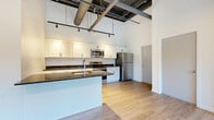Located in Rochester's vibrant East End neighborhood, this one-bedroom, one-bathroom urban loft at 50 Prince Street blends industrial character with modern finishes. Soaring ceilings with exposed ductwork and bold steel beams define the open-concept living space, giving the unit a distinctive loft aesthetic throughout. Light hardwood flooring flows seamlessly from room to room, enhancing the bright, airy feel.

The kitchen is a standout feature, offering white shaker cabinetry, dark granite countertops, a central island with a built-in sink, and a full suite of stainless steel appliances including a range, over-the-range microwave, and refrigerator. Track lighting illuminates the space with a modern, industrial touch. The kitchen opens directly to the combined dining and living area, making it ideal for entertaining.

The bedroom is generously proportioned and benefits from a dramatic bank of tall, narrow windows that flood the room with natural light and offer views of the surrounding Rochester streetscape. A frosted glass sliding door adds a sleek, contemporary element to the space.

The bathroom mirrors the kitchen's design language, featuring white cabinetry, granite countertops, a large mirror, and light hardwood-style flooring. A hallway and closet provide practical storage. This thoughtfully designed loft offers an engaging urban living experience in the heart of Rochester.