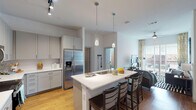 This well-appointed 4-bedroom, 4-bathroom unit in College Station, Texas offers a thoughtfully designed single-floor layout that balances comfort and functionality. The open-concept kitchen is a standout feature, showcasing grey shaker-style cabinetry, white quartz countertops, stainless steel appliances including a French door refrigerator and electric range, and a center island with a built-in sink and bar seating for multiple guests. Elegant pendant lighting and track lighting illuminate the space, while warm hardwood-style flooring flows throughout the main living areas. The kitchen connects seamlessly to the living room, which is brightened by large sliding glass doors leading to an outdoor area and featuring a ceiling fan overhead. Each bedroom offers plush carpeting, generous wall space, and access to private or semi-private bathrooms. The bathrooms are clean and modern, with white subway tile surrounds, soaking tubs, white vanities with undermount sinks, and wood-look flooring. Several bedrooms include dedicated closet space and direct sightlines to the kitchen, making this layout practical for shared living. The overall design is bright and contemporary, with high ceilings and a neutral palette throughout.