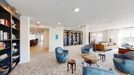 This single-floor communal amenity space offers a well-appointed and inviting environment designed for both relaxation and social engagement. The main lounge area serves as the heart of the floor, furnished with plush blue velvet swivel chairs, a rich cognac leather sofa, and an assortment of wood accent tables arranged atop a patterned, geometric carpet. Recessed lighting throughout keeps the space bright and welcoming, complemented by large windows dressed in sheer white curtains and roller shades that allow ample natural light to filter in.

A built-in floor-to-ceiling dark bookshelf unit serves as a striking architectural focal point, filled with books and decorative objects. Adjacent seating alcoves provide quieter spots for reading or conversation, while a dedicated game room features a large hexagonal card table, surrounding upholstered seating, and a wall-mounted television set against a bold geometric-tiled accent wall with under-cabinet storage.

A separate dining area with hardwood-style flooring accommodates group gatherings, visible through a connecting doorway. Wide corridors with patterned carpet and dark metal railings connect the various zones, offering glimpses into additional rooms. Curated artwork adorns the walls throughout, lending a polished, residential feel to this thoughtfully crafted communal space.