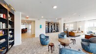 This single-floor communal amenity space offers a well-appointed and inviting environment designed for both relaxation and social engagement. The main lounge area serves as the heart of the floor, furnished with plush blue velvet swivel chairs, a rich cognac leather sofa, and an assortment of wood accent tables arranged atop a patterned, geometric carpet. Recessed lighting throughout keeps the space bright and welcoming, complemented by large windows dressed in sheer white curtains and roller shades that allow ample natural light to filter in.

A built-in floor-to-ceiling dark bookshelf unit serves as a striking architectural focal point, filled with books and decorative objects. Adjacent seating alcoves provide quieter spots for reading or conversation, while a dedicated game room features a large hexagonal card table, surrounding upholstered seating, and a wall-mounted television set against a bold geometric-tiled accent wall with under-cabinet storage.

A separate dining area with hardwood-style flooring accommodates group gatherings, visible through a connecting doorway. Wide corridors with patterned carpet and dark metal railings connect the various zones, offering glimpses into additional rooms. Curated artwork adorns the walls throughout, lending a polished, residential feel to this thoughtfully crafted communal space.