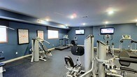 This single-level fitness center offers a functional and focused workout environment designed to accommodate a range of exercise needs. The room is finished with deep blue-painted walls and a low ceiling outfitted with recessed lighting, creating a focused, energetic atmosphere well-suited for exercise. Rubber flooring throughout provides both durability and safety underfoot.

The space is equipped with a solid selection of fitness equipment, including multiple strength-training machines positioned toward the front and sides of the room, offering users options for resistance and muscle-targeted workouts. A treadmill and stationary bike are positioned toward the rear, catering to cardio-focused routines. Two wall-mounted flat-screen televisions are installed at different points along the walls, allowing users to stay entertained during workouts. A whiteboard and a framed motivational print add subtle decorative and functional touches to the walls. Ventilation is provided via ceiling vents, and an illuminated exit sign ensures clear safety guidance. The layout is efficient, making good use of the available floor space while keeping equipment accessible and unobstructed.
