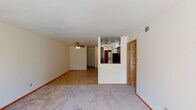 This single-floor residential unit offers a functional and straightforward layout designed for comfortable everyday living. The open living area is carpeted and features a ceiling fan with light fixture, providing both comfort and airflow. A distinctive half-wall partition separates the living space from the kitchen, creating visual openness while defining each zone clearly. The kitchen is well-equipped with warm wood cabinetry, dark laminate countertops, a black electric range with overhead ventilation, and a stainless steel sink, all set over tile flooring. Natural light filters into the living area through a sliding glass door fitted with vertical blinds, offering views of a grassy outdoor area. An additional bedroom features a large window with neighborhood views and plush carpeting, making it a bright and comfortable retreat. Sliding panel closets provide practical storage in the bedrooms. The bathroom includes a wide vanity with a white countertop, wood cabinetry, a lighted mirror, and tile flooring, alongside a separate tub and shower combination surrounded by clean white subway tile. Warm wood-toned door and trim details are consistent throughout, lending a cohesive and classic character to the entire space.