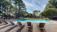 Located at 101 Royal Dr in Madison, Alabama, this two-floor community offers a well-appointed array of outdoor amenities set against a backdrop of tall, mature pine trees. The centerpiece of the outdoor area is a generously sized swimming pool with crystal-clear blue water, accessible via entry steps and surrounded by a wide concrete pool deck. Numerous lounge chairs are arranged poolside, providing ample space for relaxation under open skies. A built-in brick fire pit is positioned near the deck, adding a welcoming gathering spot for cooler evenings. The community also features a covered outdoor grill station with seating and a shaded umbrella table, ideal for outdoor dining and socializing. A dedicated playground with a slide is visible within the fenced grounds, catering to younger residents. The clubhouse building is a standout architectural feature, showcasing a gabled roofline, covered front porch with seating, arched entryway doors, and a chimney — all framed by manicured landscaping. The entire outdoor amenity area is enclosed by a black iron fence, offering both security and an open, airy feel. The lush green surroundings and towering pines lend the space a serene, park-like atmosphere throughout.
