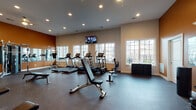 Located at 12509 Crown Park Ct in Charlotte, NC, this single-floor fitness center offers a comprehensive workout environment designed to accommodate a wide range of exercise needs. The space is defined by its open layout, durable rubber flooring, and a high ceiling with recessed lighting that keeps the area bright and functional throughout the day.

A bold terracotta accent wall anchors one end of the room, adding visual warmth and contrast against the neutral beige walls. Large multi-pane windows line the perimeter, inviting substantial natural light and providing views of the surrounding neighborhood. Wall-mounted flat-screen televisions are positioned at multiple points for easy viewing during workouts.

The cardio zone is well-appointed with a row of Landice treadmills and elliptical machines positioned along the windowed wall. The strength training area features a cable crossover machine, multiple selectorized weight machines, a dedicated free weights section with a full dumbbell rack, and adjustable benches. Additional equipment includes stability balls, medicine balls, and exercise mats stored along the accent wall. Full-length mirrors along one wall enhance spatial awareness and allow users to monitor their form. The overall atmosphere is energetic yet practical, making this fitness center a thorough and accessible facility.
