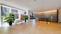 This expansive residential lobby offers a refined first impression through a thoughtfully designed, modern interior. Upon entry, large-format stone and light wood tile flooring transitions seamlessly across zones, anchoring the open layout. A dramatic open-riser staircase with warm wood treads, steel framing, and glass railings serves as a striking architectural focal point, rising beneath soaring double-height ceilings framed by floor-to-ceiling glazing that floods the space with natural light and frames views of a lush landscaped garden courtyard beyond. A sleek reception desk, warmly lit from below and backed by a textured vertical wood slat wall with integrated accent lighting, provides an elegant welcome point. The lounge area features a linear fireplace set into a stone surround, accompanied by contemporary seating arrangements that invite residents to gather in comfort. Curated greenery—including large potted tropical plants—adds life and warmth throughout. A dedicated corridor leads to a bank of elevators and a built-in mailroom, while decorative convex mirrors and carefully selected artwork adorn the walls, contributing to the gallery-like atmosphere. The overall ambiance is one of calm sophistication, blending natural materials, architectural detail, and abundant light into a cohesive, welcoming environment.