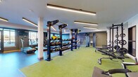 Located at 705 First Ave SW in Rochester, Minnesota, this well-appointed fitness center delivers a comprehensive workout environment within a single, open-plan floor. The space is thoughtfully divided into distinct functional zones, each designed to support a variety of training disciplines.

The cardio area lines the perimeter near large windows, housing a row of Star Trac treadmills, elliptical trainers, and stair climbers that benefit from abundant natural light and outdoor views — an energizing backdrop for endurance training. The flooring in this zone features wood-effect paneling, lending a clean, modern aesthetic.

At the heart of the facility, vibrant green turf flooring defines the strength and functional training area. Here, Octagon multi-functional rigs serve as the centerpiece, supporting pull-up bars, cable attachments, and TRX suspension trainers. Tiered racks are fully stocked with medicine balls, slam balls, and kettlebells in varied weights, providing versatile options for dynamic workouts.

A dedicated free-weight and strength zone features TAG power racks, barbells, and adjustable benches positioned on turf, ideal for compound lifting. A cable machine and additional accessories round out the strength offering.

Overhead, linear LED lighting fixtures illuminate the entire space evenly, complementing the contemporary design. Accent walls in deep blue-gray tones add visual depth, while structural columns are cleanly integrated throughout. A water station is conveniently located within the facility.