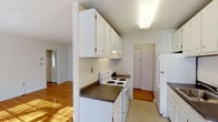 This single-floor unit in Versailles offers a clean, functional layout with consistent design details throughout. Warm hardwood flooring runs through the main living and sleeping areas, reflecting natural light and adding a sense of warmth to the bright, white-walled interiors.

The living room is generously proportioned, featuring a ceiling fan, baseboard heating, and a bank of windows that allows abundant natural light to pour in. The open feel of the space makes it highly versatile for various furniture arrangements.

A separate bedroom provides a quiet retreat, also finished with the same rich hardwood floors. A window fitted with blinds and a wall-mounted air conditioning unit ensures year-round comfort.

The galley-style kitchen is efficiently designed with white cabinetry lining both walls, offering ample upper and lower storage. Dark laminate countertops provide a practical contrast, while a white electric range, refrigerator, and stainless steel sink complete the functional setup. Light tile flooring ties the kitchen together, and a doorway leads to additional utility space beyond.

The bathroom is finished with light tile flooring and features a bathtub with shower, a toilet, a window for natural ventilation, and a louvered storage closet. Baseboard heating and a grab bar add practical convenience. The overall design of this unit is straightforward and move-in ready.