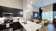 This single-story residence offers a well-appointed 1-bedroom, 1-bathroom layout designed with modern finishes and an open, airy feel. Dramatically high ceilings define the main living and kitchen areas, amplifying the sense of space and allowing natural light to flow freely through large windows dressed with floor-length drapes.

The kitchen is outfitted with dark shaker-style cabinetry extending toward the ceiling for ample storage, stainless steel appliances including a French door refrigerator, electric range, and built-in microwave, as well as a white quartz countertop that extends into a peninsula with bar seating. A classic white subway tile backsplash and globe pendant lighting add refined detail.

The open living area benefits from soaring ceilings and a ceiling fan, creating a comfortable, well-ventilated space. The bedroom features plush carpeting, a window with natural light, and elevated ceiling height. An in-unit laundry area is conveniently located near the bathroom.

The bathroom showcases dark vanity cabinetry paired with a white countertop and a full tub and shower surrounded by large-format marble-look tile, creating a clean, spa-like aesthetic throughout.