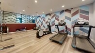 Located at 5095 E Donald Ave in Denver, Colorado, this combined gym and laundry facility offers a thoughtfully designed space that serves both fitness and practical needs. The fitness area is visually striking, featuring a bold chevron-patterned accent wall in shades of blue, burgundy, and white that anchors the room with a modern, energetic aesthetic. Complementing this is a deep crimson lower accent wall, topped by a decorative metal chevron partition that adds architectural depth and visual separation within the space. Light wood-look flooring runs throughout, contributing to a clean and contemporary feel. Recessed lighting illuminates the gym evenly, creating a bright and inviting environment. The fitness area is equipped with a range of cardio machines including treadmills and an elliptical, a multi-function cable machine, a dumbbell rack with free weights, a weight bench, a stationary bike, and exercise balls. The laundry room, visible in the floorplan, is outfitted with multiple washers and dryers arranged for convenient side-by-side use. The facility is accessed via glass-paneled doors and includes clearly marked emergency exits, ensuring safety throughout. Overall, this space combines functional amenities with polished, modern interior design.