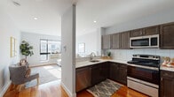 Located at 697 Bronx River Rd in Yonkers, NY, this single-floor residence offers a thoughtfully designed layout that balances modern finishes with comfortable living. Rich hardwood flooring flows throughout the main living areas, creating a warm and cohesive aesthetic from entry to living room.

The kitchen is outfitted with dark espresso cabinetry, white subway tile backsplash, light countertops, and a full suite of stainless steel appliances including a gas range, built-in microwave, dishwasher, and refrigerator. A half-wall separates the kitchen from the living area, preserving an open, connected feel while providing a sense of defined space. The living room benefits from large windows that invite generous natural light and offer views of the surrounding neighborhood.

The bedroom is a comfortable retreat, featuring a sizable window with exterior views, hardwood flooring, and soft neutral wall tones that create a calm atmosphere. The bathroom is finished with white subway tile wainscoting, marble-look tile flooring, a white vanity with gold-toned fixtures, and a glass-enclosed soaking tub and shower combination, lending a clean and polished look. The entry area provides a practical transition point with direct access to the kitchen. Overall, the residence presents a modern, well-appointed interior with consistent finishes throughout.