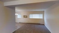 This single-floor space features an open main living area finished with wall-to-wall carpeting in a neutral tone, contributing to a quiet and functional atmosphere. Two windows fitted with vertical blinds allow natural light to fill the room, enhancing the sense of openness. A wall-mounted air conditioning unit provides climate control, complemented by baseboard heating that runs along the perimeter of the room for year-round comfort. A pendant light fixture with multiple bulbs is visible, offering overhead illumination. The walls are finished in a clean, neutral off-white tone, and the space features a structural beam detail overhead that adds subtle architectural definition. The layout appears straightforward and practical, with an open flow that allows for flexible furniture arrangement. Overall, the space presents a simple, utilitarian design with essential comfort features in place.