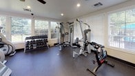 This fitness center is a comprehensively outfitted single-floor exercise facility designed to support a full range of workout routines. The space is lined with large windows along multiple walls, flooding the interior with abundant natural light and offering pleasant views of the surrounding greenery — creating an inviting and energizing atmosphere for exercise.

The flooring throughout is durable rubber gym matting, providing both safety and comfort underfoot. A ceiling fan keeps air circulating, complemented by recessed lighting and HVAC vents for climate control year-round. A security camera is also present, enhancing safety within the space.

The cardio section features multiple Matrix-brand treadmills and an elliptical trainer, ideal for endurance and aerobic workouts. A rowing or cycling machine adds further variety to cardiovascular training options. For strength training, the facility includes a multi-station cable machine by Hoist, a dedicated weight bench, and a full rack of dumbbells spanning a wide range of weights — accommodating both beginners and experienced users.

Full-length wall mirrors on one side of the room visually expand the space and allow users to monitor their form during exercise. Wall-mounted televisions provide entertainment during workouts. The entrance features elegant frosted-glass double doors, lending a polished, professional finish to this thoughtfully designed fitness facility.