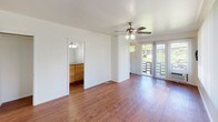 Located in Moana Vista in the heart of Honolulu, Hawaii, this single-floor residence offers a well-organized and functional layout with a warm, understated aesthetic throughout. Rich hardwood flooring runs continuously across the living spaces, lending a cohesive and inviting feel to the entire unit.

The living area is generously proportioned, bathed in natural light streaming through large windows dressed with vertical blinds. A ceiling fan provides comfortable airflow, complementing the bright and airy atmosphere. The room offers versatile open space suitable for a variety of furniture arrangements.

Adjacent to the living area, the kitchen is efficiently designed with warm wood cabinetry providing ample storage, a gas range, a full-size refrigerator, and a countertop sink. The layout maximizes every inch of available space while maintaining a clean and practical cooking environment.

A vanity area with wooden cabinetry and a large mirror is accessible from the main living space, offering additional storage and convenience. The bathroom is cleanly finished in white tones, featuring a single-basin vanity with a large mirror and a tiled walk-in shower with a natural light window.

An outdoor balcony, accessible through sliding glass doors, brings a touch of Hawaii's lush greenery indoors, enhancing the overall connection to the tropical surroundings of Honolulu.
